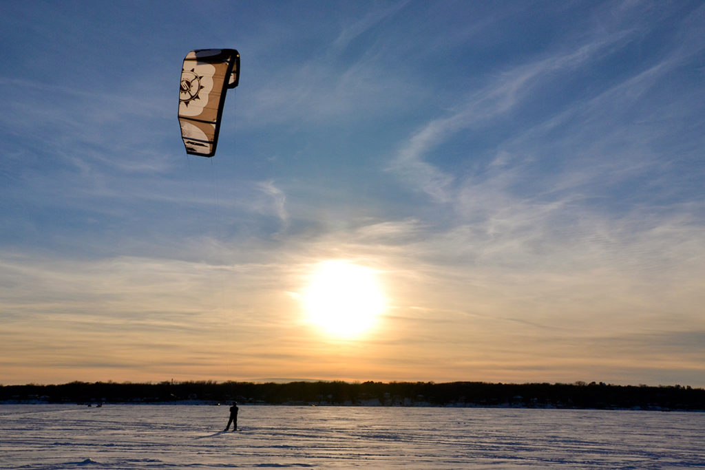 A kiteboarder rides across a frozen lake on his snowboard with the sunset in the background.